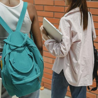 Back view crop female students wearing casual wear with backpacks and textbooks strolling together on sidewalk against brick wall and chatting in university campus
