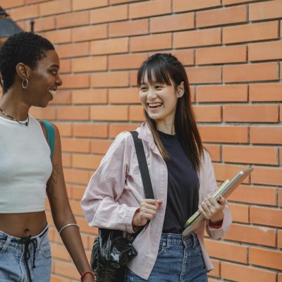Cheerful multiracial female students wearing casual clothes carrying backpack and notebooks near college brick wall while discussing plans and looking at each other with smiles