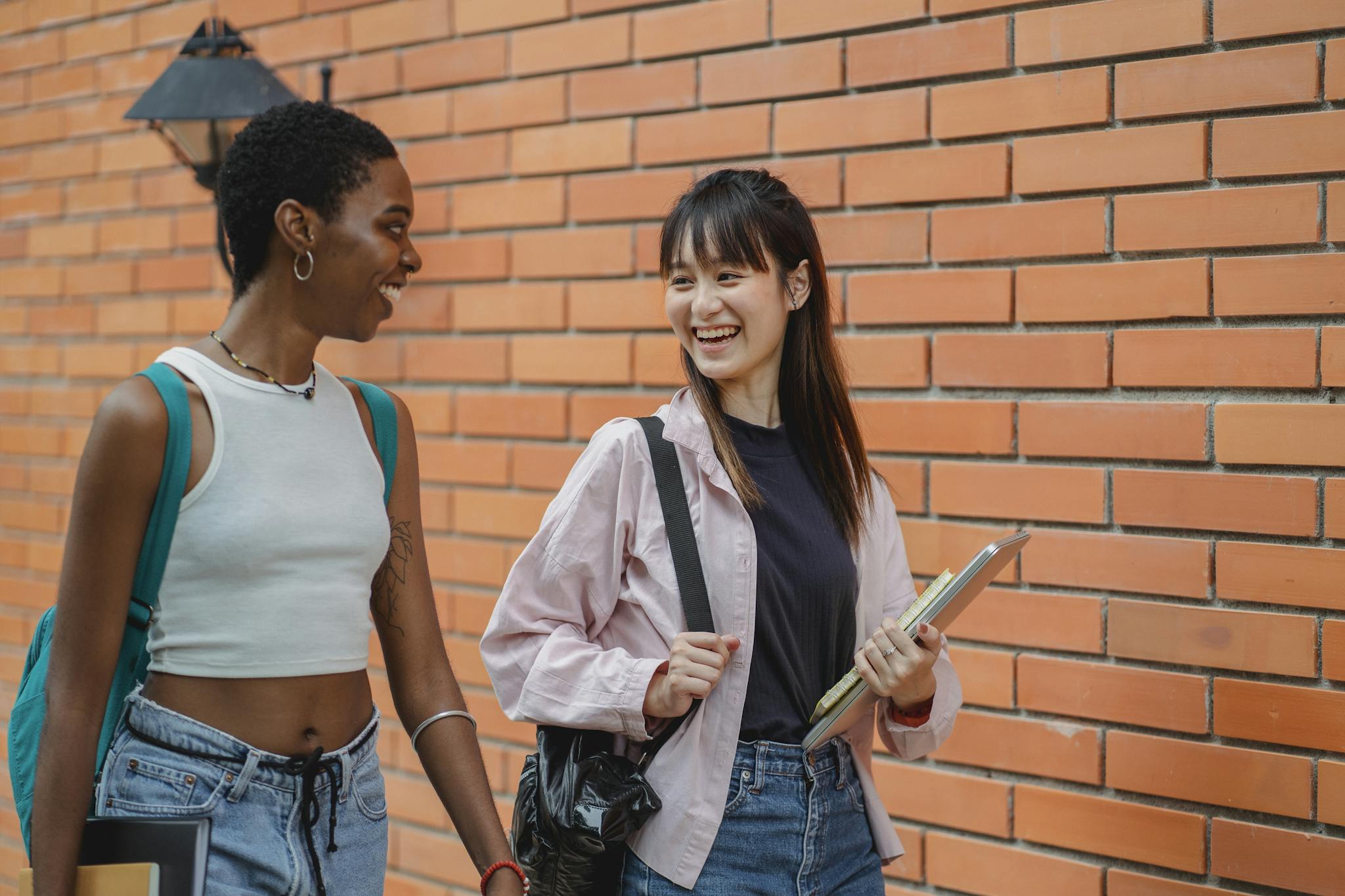 Cheerful multiracial female students wearing casual clothes carrying backpack and notebooks near college brick wall while discussing plans and looking at each other with smiles