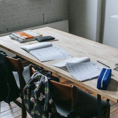 Wooden desk with opened copybooks and workbooks placed near chairs with backpacks in light classroom